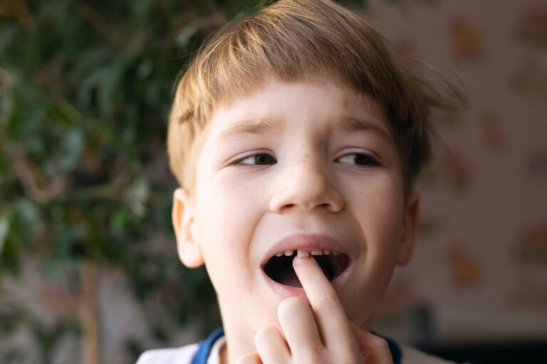 A child with short hair touches a gap in their teeth, likely where a tooth is missing. A green plant is visible in the blurred background.
