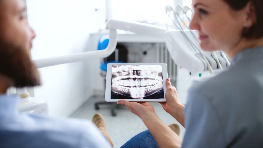 A dentist shows a patient a digital dental x-ray image on a tablet in a bright, modern dental office.