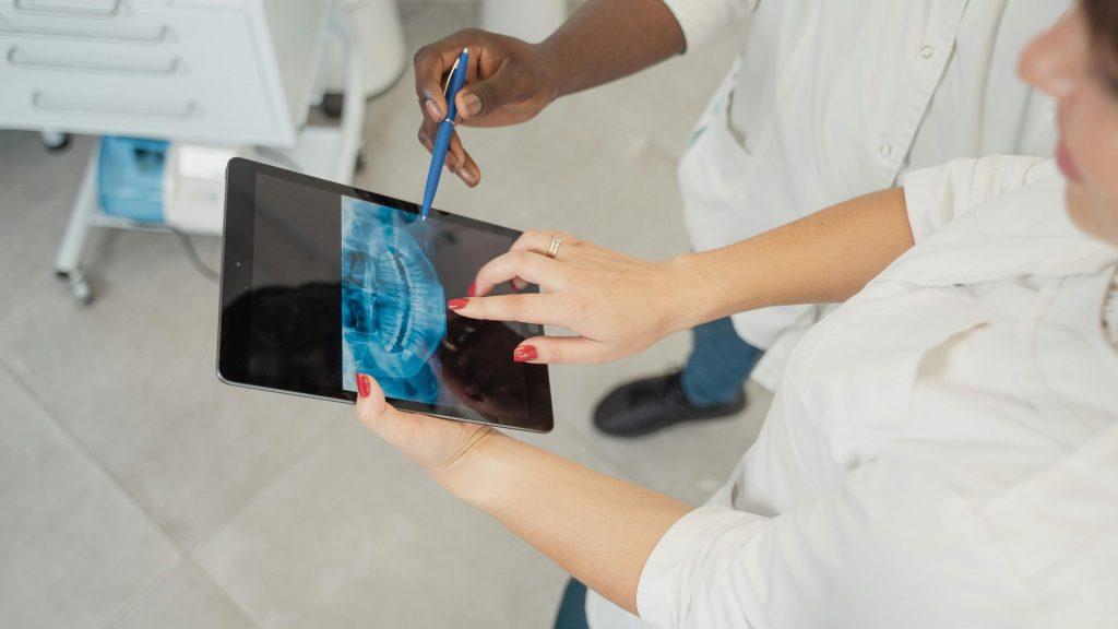 Two people examine a digital dental x-ray on a tablet; one points at the x-ray image with a pen while the other gestures on the screen.