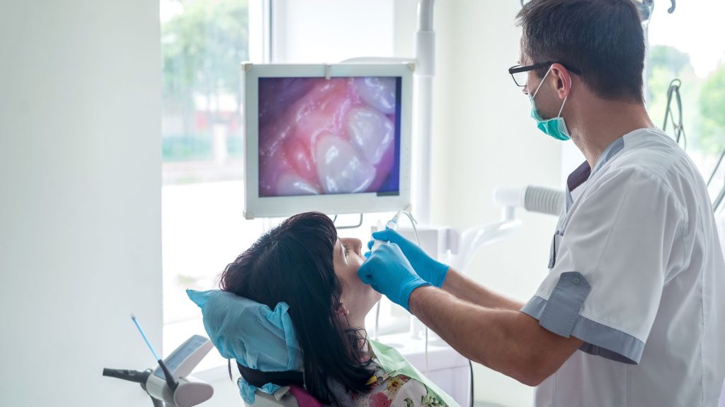A dentist examines a patient's mouth while an intraoral camera and digital dental x-rays display detailed images of teeth and gums on a monitor.
