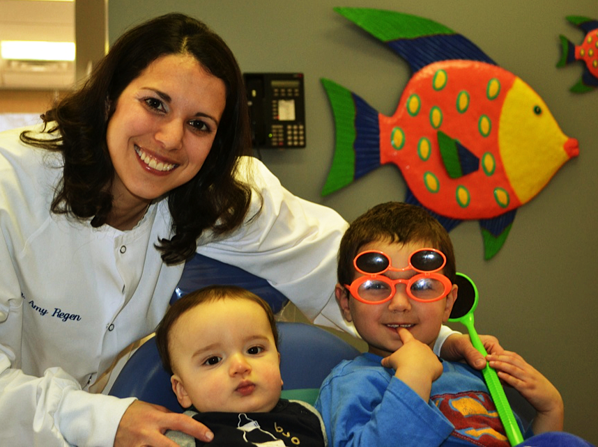 A woman in a white coat poses with two young children; one child wears sunglasses with multiple lenses. A colorful fish decoration is on the wall behind them.