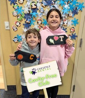 Two smiling children hold decorated pencil cases and a "Cavity-Free Club" sign in front of a wall with paper snowflakes and cartoon faces.