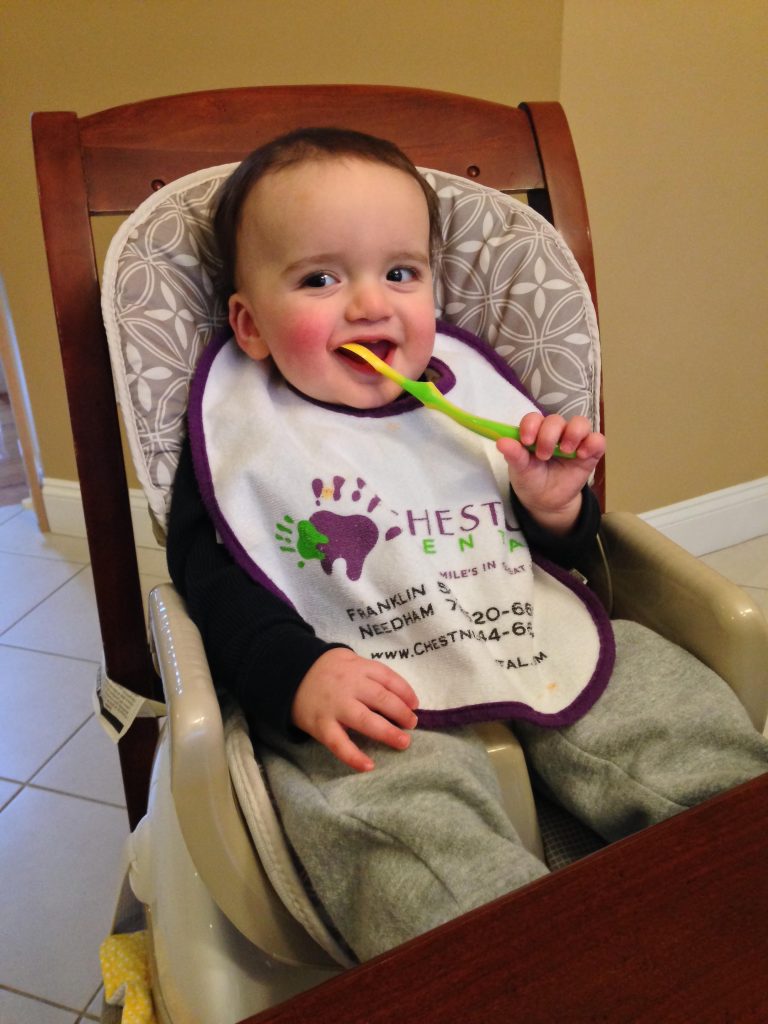 A toddler sits in a high chair wearing a bib and holding a green and orange toothbrush to their mouth, smiling.