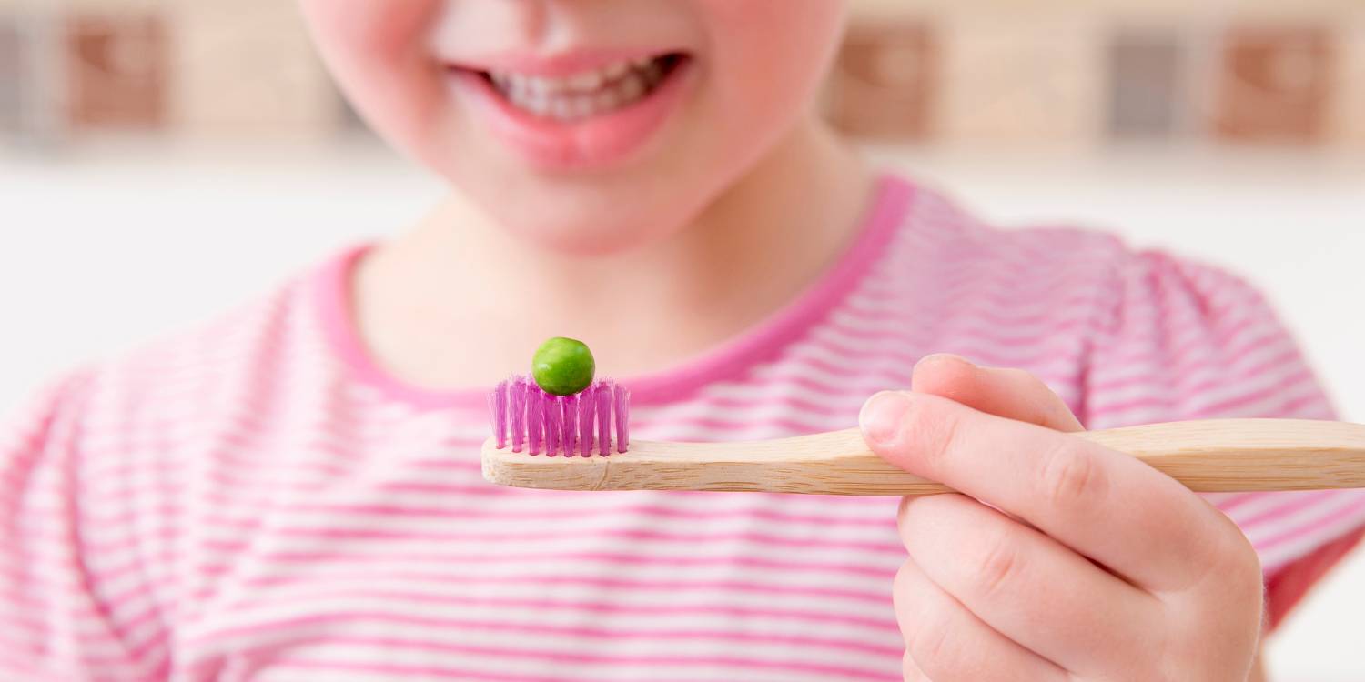 Selective focus on bamboo tooth brush with green pea on it and smiling blurred anonymous girl on background. Children should use the size of a pea amount fluoride toothpaste on brush concept.
